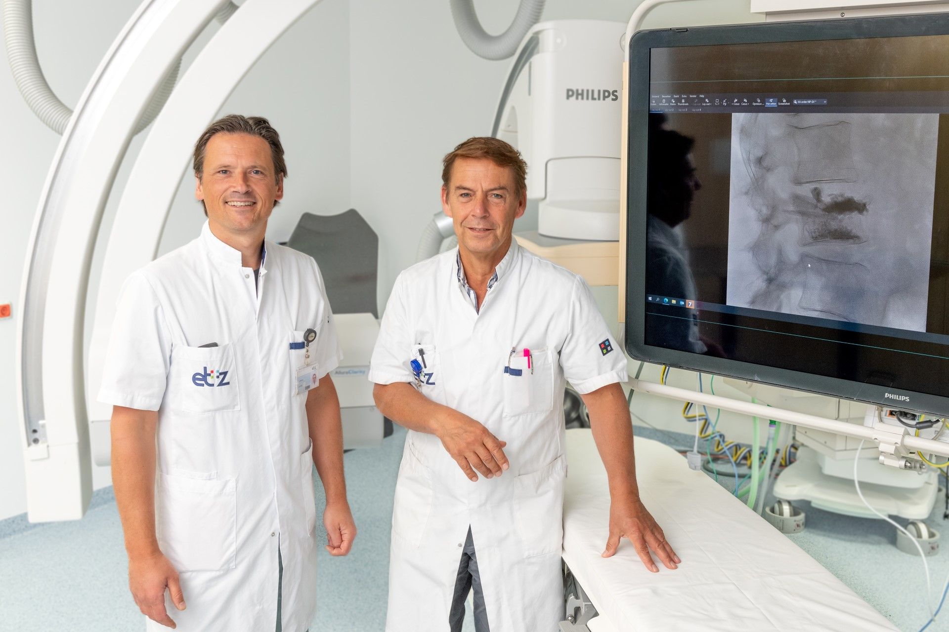 dr. Venmans en dr. Lohle staan in de intervieruimte. Op het beeldscherm staat een röntgenfoto van een ruggenwervel, donkerdere vlekken tonen het ingespoten botcement aan. Slingers versieren de apparatuur feestelijk.