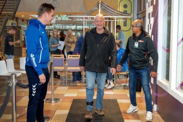 Een man loopt over een zwarte mat bestaande uit veel losse vlakken. Hij wordt begeleid door 2 sport- en beweegcoaches.
