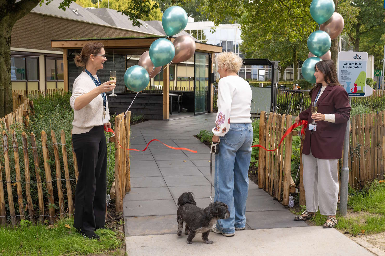 De houten omheining van de groene huisdierentuin is versierd met ballonnen en een rood lint. Het lint wordt doorgeknipt.